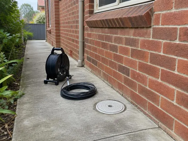 Capped drain inspection opening beside a coiled camera reel on a concrete side path by a brick house in Normanhurst