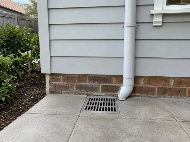 Outdoor grated gully beside a brick and weatherboard house with dry concrete paving in Asquith