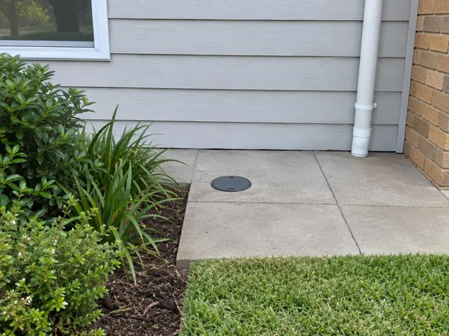 Capped access point in a concrete patio beside a house wall and garden bed in Berowra